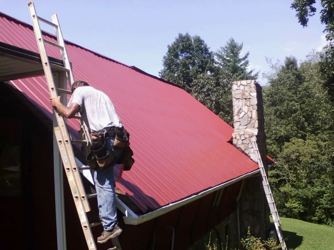 Roofer on a ladder beside a red metal roof and stone chimney — Soffit & Fascia Repair by Roofing Brothers in Egg Harbor Township